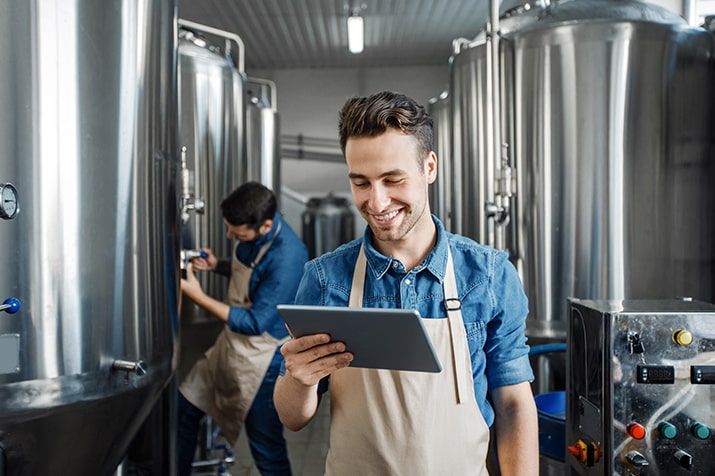 Aplicacoes-checklist-manutencao-industrial-min - Blog SULTS Manutenção industrial - Homem jovem com tablet na mão, conferindo informação dentro da indústria de cervejas.