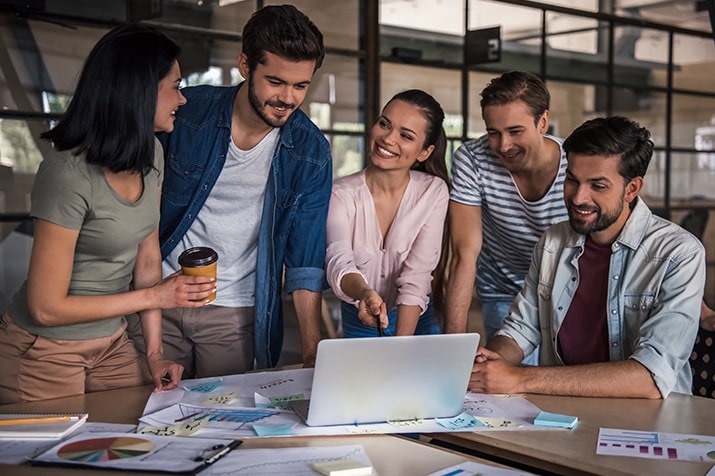 Conceito-inovacao-empresarial-SULTS-min - Blog SULTS Inovação empresarial - grupo de colaboradores jovens ao redor da mesa de escritório olhando para notebook sorrindo.