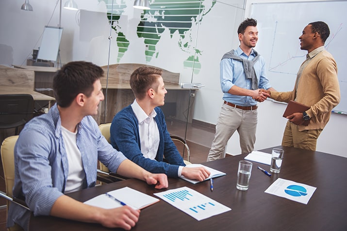 Dicas-para-comunicacao-assertiva-SULTS-min - Blog SULTS Comunicação assertiva - Reunião de colaboradores homens na empresa. Dois estão sentados sorrindo e olhando para os dois em pé, que estão se cumprimentando.