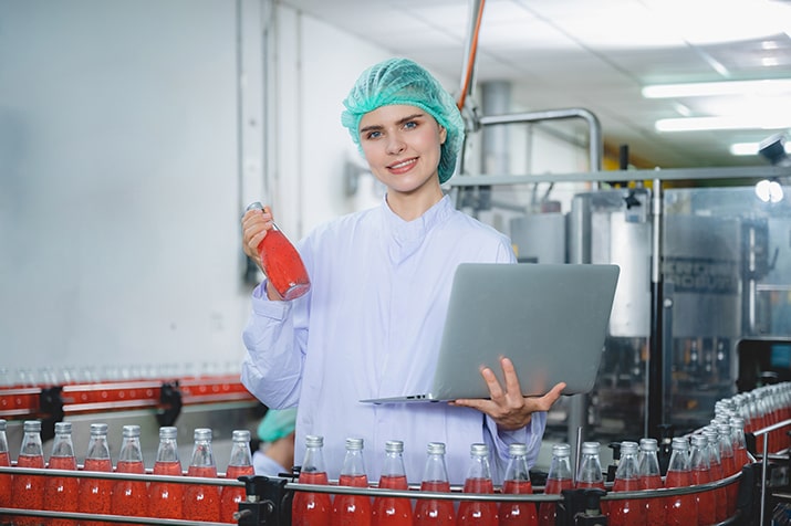 Tipos-de-checklist-redes-de-alimentacao-SULTS-min - Blog SULTS Tipos de checklist - Operadora de fábrica de bebidas segurando frasco cheio e um notebook, sorrindo para a câmera.