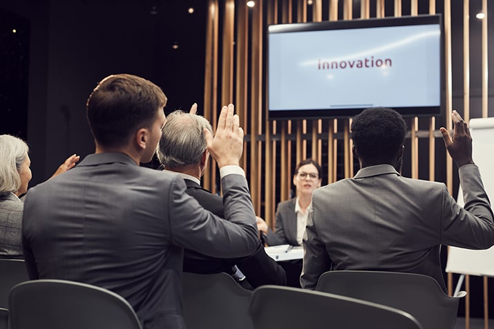 Tipos-de-inovacao-empresarial-SULTS-min - Blog SULTS Inovação empresarial - colaboradores reunidos assistindo à palestra sobre inovação com o braço levantado para tirar dúvidas.