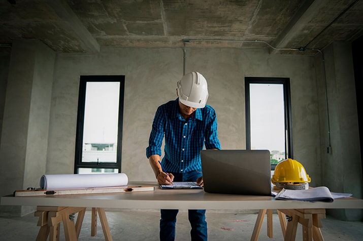 Construtechs-SULTS - Blog SULTS Construtechs - trabalhador em obra conferindo planta da casa com notebook ao lado, sobre a mesa.
