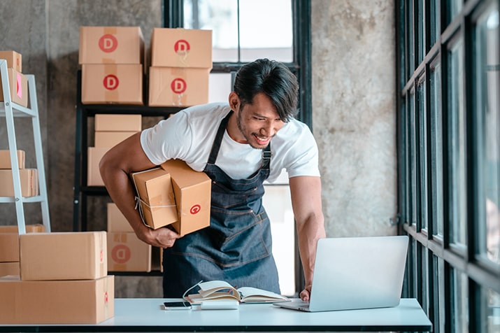 O-e-commerce-SULTS - Blog SULTS E-commerce - colaborador de loja carregando caixas e sorrindo para laptop.